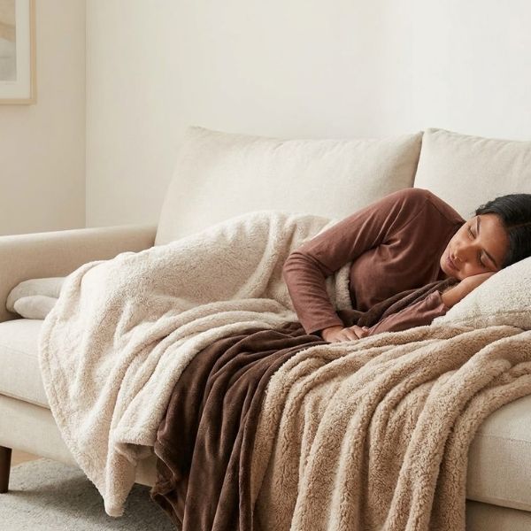 An individual relaxes comfortably on a plush sofa, wrapped in soft, knitted blankets.