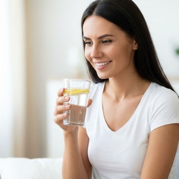 A healthy woman smiles gently while holding a glass of lemon water in a clean, softly lit room, showing fully healed skin.