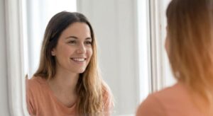 A smiling woman looks closely at her reflection in a large, sunlit mirror.